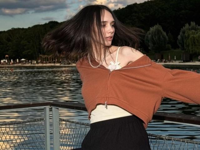 SherryMalara, avec de longs cheveux bruns, un pull marron à une épaule, un crop top blanc et une jupe noire, se tient près d'un cours d'eau réfléchissant, en train de tourner, et montre légèrement ses seins..