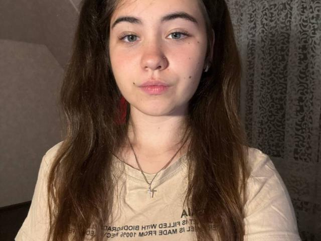 LeliaSixon tem cabelos castanhos longos, pele clara e olhos azuis, com uma pequena pinta acima dos lábios rosados. Ela está vestindo uma camisa bege com um colar de cruz..
