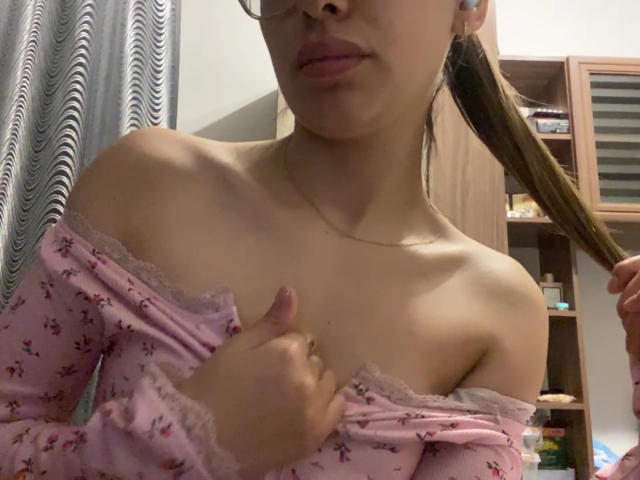 RaneeDerienzo with light skin, small tits, wearing pink off-shoulder floral top, holding hair, glasses, subtle makeup, small necklace, right hand touching chest, left hand holding hair.