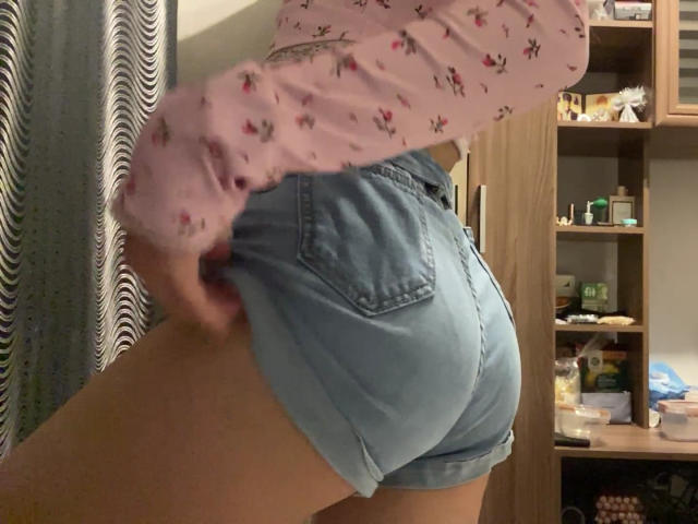 RaneeDerienzo trägt kurze, blaue Jeansshorts, die ihr Hintern betonen, und ein pinkes Langarmshirt mit kleinen roten Mustern. Sie zieht die Shorts leicht hoch, um mehr von ihrem Hintern zu zeigen..
