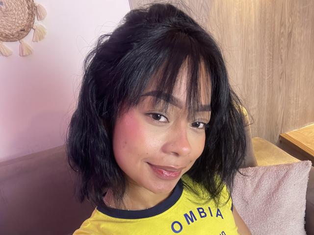 AbbieTaylors has messy black hair, dark eyebrows, pink cheeks, and a small beauty mark on her right cheek. She's wearing a yellow shirt, smiling slightly.