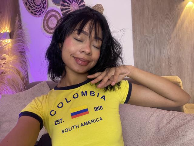 AbbieTaylors with closed eyes, smiling, wearing a yellow Colombia t-shirt, has black hair, and is touching her cheek. Her tits are small and her skin is tan.