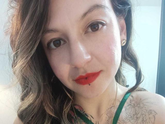 Ariana: Loira com batom vermelho, olhos castanhos e cabelo castanho ondulado, tem um piercing no lábio, uma blusa verde com tiras e uma tatuagem floral no ombro. Ela está usando brincos pequenos..