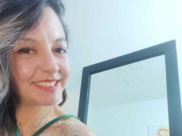 ArianaBlonde hat ein Lächeln mit einem Lippenpiercing, einen grünen Gürtel, ein Tattoo auf der Schulter und welliges, schwarz-graues Haar. Im Hintergrund ist ein Spiegel zu sehen. Helle Haut, braune Augen. Oberkörper-Selfie..