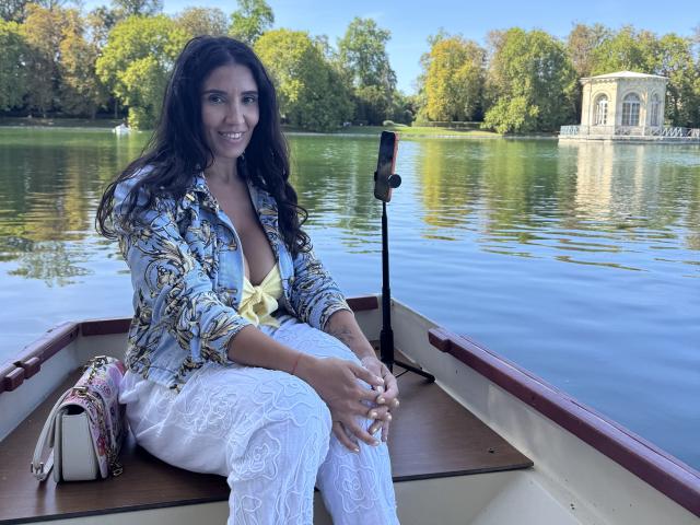 Samia Khalifa, con cabello largo y oscuro, vestida con una chaqueta azul estampada, una blusa amarilla y pantalones blancos de encaje, sentada en un bote, sonriendo, con un bolso pequeño en el asiento..