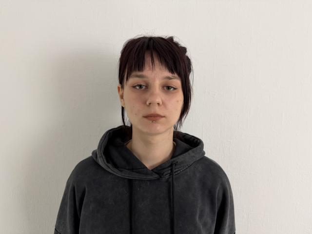 TeshaDotter tiene una expresión apagada, cabello lacio y oscuro con flequillo, una sudadera gris y un pequeño piercing en la nariz. Está de pie frente a una pared blanca, mirando directamente a la cámara..
