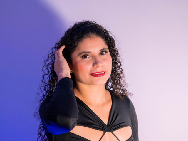 Katteryn Soto, con cabello negro rizado, lápiz labial rojo y un top negro con aberturas, con la mano en el cabello, sonriendo, piel marrón clara, senos pequeños, atractiva, en una foto bien iluminada con tonos azules..