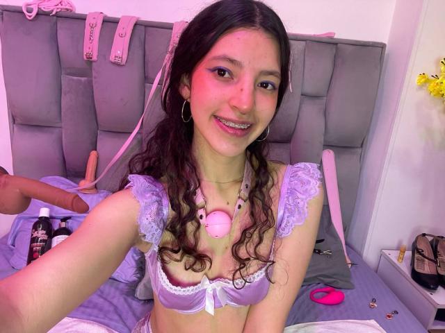 NamySex, avec ses cheveux bruns bouclés, ses joues roses et son soutien-gorge en dentelle violette, souriant à la caméra, portant un bouchon buccal rose, sur un lit avec un stimulateur et des jouets sexuels..