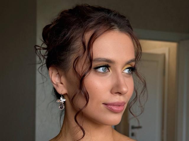 ShalonGluc has wavy brown hair, blue eyes, and pink lipstick, wearing purple earrings with a floral design, looking to the side with a slight smile.