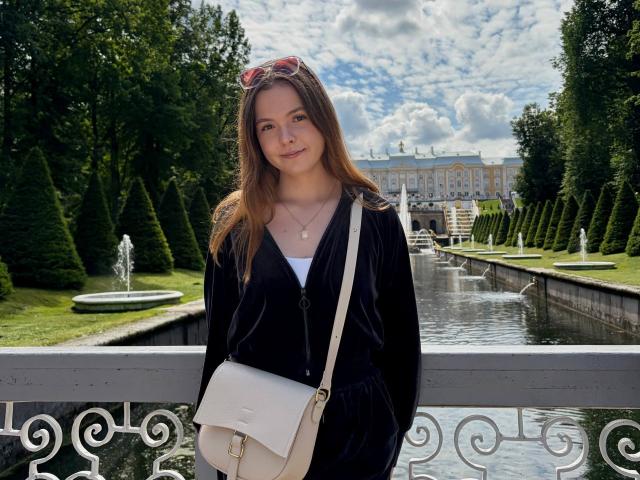 JeseniaPiontek with brown hair, black velvet top, white cross necklace, pink sunglasses on head, white purse, standing by ornate white railing, water fountain in background, cloudy sky.