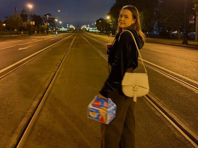 JeseniaPiontek stands on a dark street, back to camera, holding a blue box and white bag, wearing a black velvet top and dark pants, with long brown hair.