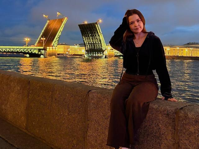 JeseniaPiontek with brown hair, black zip-up jacket, and brown pants, sitting on a concrete wall, hand in hair, bridge with green lights and water in the background.