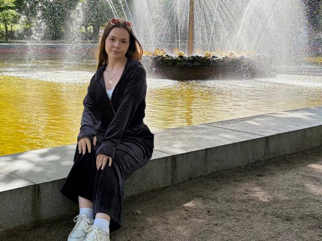 JeseniaPiontek sits on a concrete ledge wearing a black velvety zip-up dress, white sneakers, and sunglasses on her head, with water spraying from a fountain behind her.