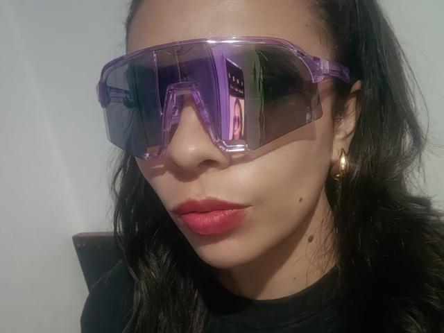 Het gezicht van VioletaStone, met paarse reflecterende brillen, rode lippenstift en zwart golvend haar, draagt gouden oorringen, een zwart shirt en een kleine moedervlek onder haar rechterwang..