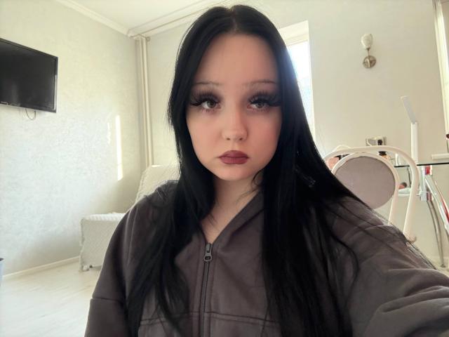 Staci Brookshear, avec ses longs cheveux noirs, son rouge à lèvres foncé et ses longs cils, porte une veste zippée grise et regarde directement la caméra dans une pièce lumineuse..