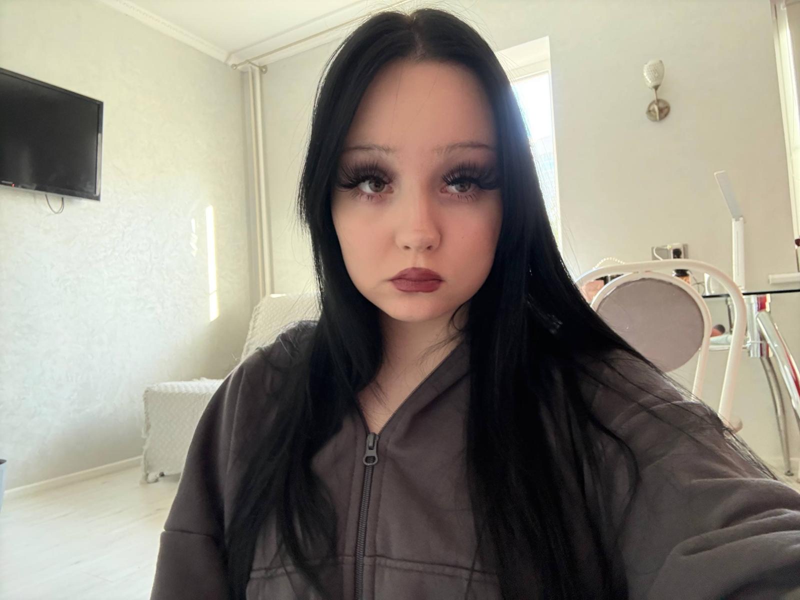 Staci Brookshear, con cabello negro largo, lápiz labial oscuro y pestañas gruesas, vestida con una chaqueta gris con cremallera, mirando directamente a la cámara en una habitación luminosa..