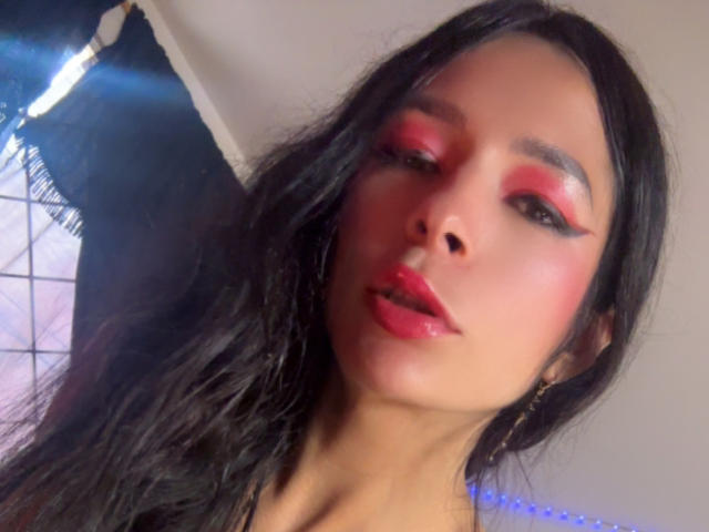 ArielNitzche with glossy pink lipstick, red eye makeup, and black wavy hair, looking at the camera with slightly parted pink glossy lips. Her face is centered in the shot.