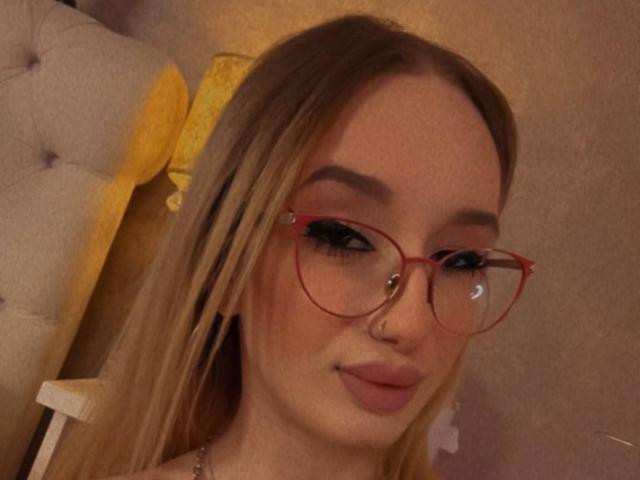 EmmaNaughtines with blonde hair, red glasses, nose ring, and pink lipstick, smiling softly. She has full eyebrows and a subtle makeup look, sitting in a cozy, dimly-lit room.