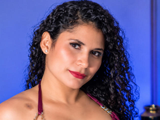 Katteryn Soto tiene el pelo negro y rizado, piel marrón media, lápiz labial rojo, pequeños aretes de diamantes y un top de tirantes color burdeos. Sus senos son parcialmente visibles. Está mirando a la cámara..