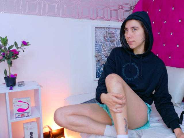 FreyaraVenwo está sentada em uma cama com as pernas cruzadas, vestindo um moletom preto, shorts verdes e meias brancas, com uma expressão séria, e atrás dela há uma parede rosa com textura..