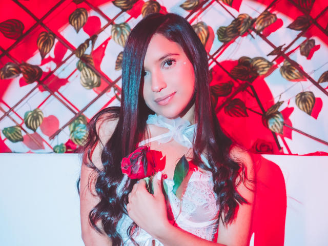 LexieJones with long brown hair, holding a red rose, wearing a white lace top, smiling at the camera, red and pink lighting, delicate white ribbon around neck.