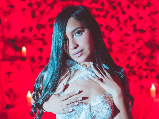 LexieJones with long black hair, white lace top, hands on her tits, red glowing background, nails painted white, looking at camera, candles in background.