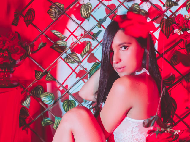 LexieJones with long black hair, red flower in hair, white lace top, red background, looking over shoulder, sitting with knees up, red and green vines in diamond pattern.