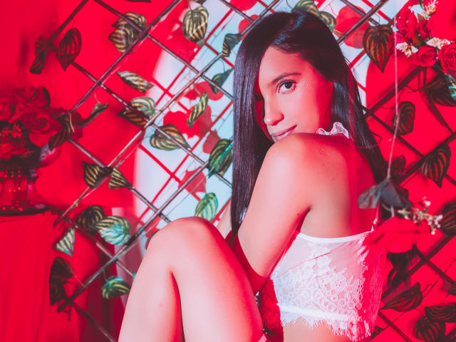 LexieJones with long black hair, looking over her shoulder, wearing white lace lingerie, sitting with knees up, red and floral background, sexy, cute, attractive, soft red lighting.