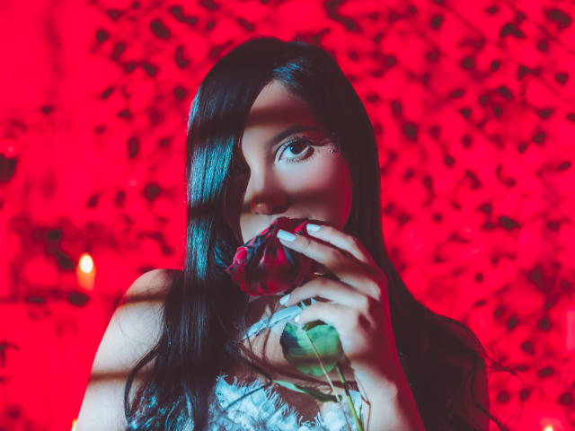 LexieJones with long black hair, holding a rose to her mouth, red light casting shadows on her face, her eye visible, looking seductive, wearing a lace top.