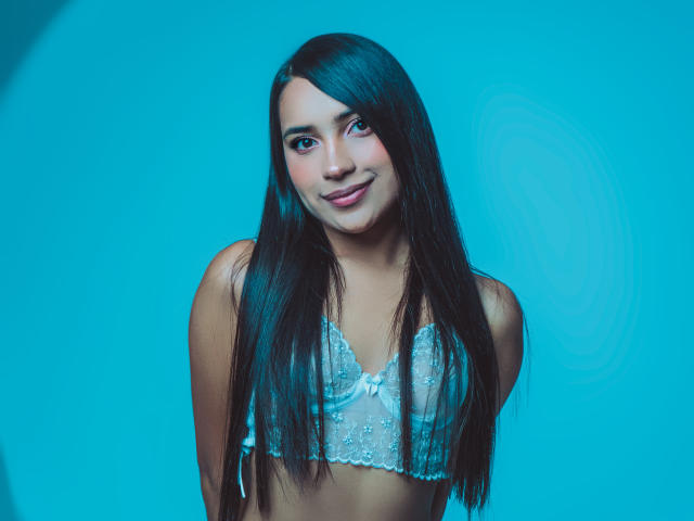 LexieJones has long, straight black hair, wearing a sheer, blue-ribboned bralette, smiling with her tits slightly visible, against a solid blue background.