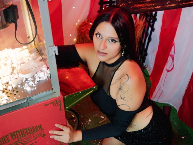 RedMoont with dark hair, blue eyes, black mesh top, and black sequin skirt, leaning on a red and green machine, showing tattoos on her arm, looking at the camera.