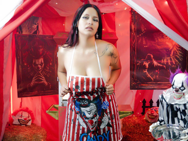 RedMoont with dark hair, large tits, and tattoos wearing a red and white striped apron with a clown design, standing in a spooky setup with horror decorations.