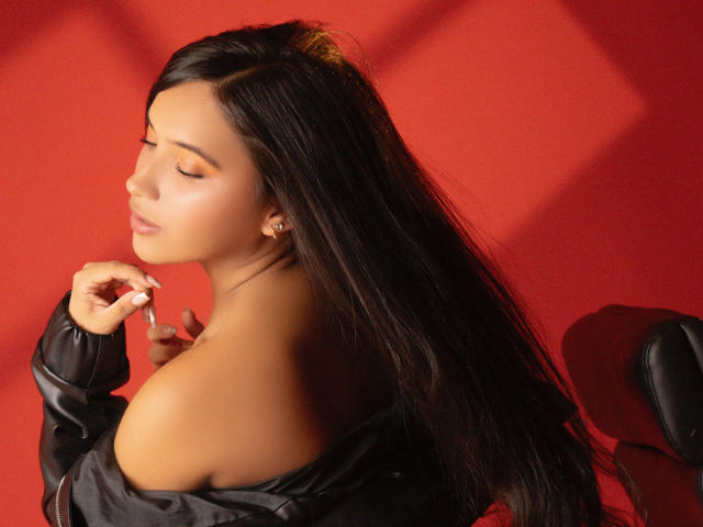 LexieJones with long black hair, eyes closed, wearing a black off-shoulder top, holding a cigarette, red background, sexy, soft light, slightly turned profile, elegant, confident.