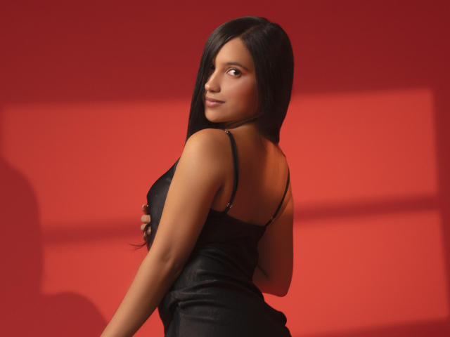 LexieJones with long black hair, wearing a black dress, looking back over her shoulder, showing her tits, against a red background, with shadows on the wall.