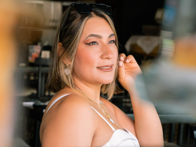 EmaFoxy, con cabello rubio hasta los hombros, una blusa blanca, un collar dorado y gafas de sol en la cabeza, sonriendo, tocándose la mejilla, con sombra de ojos rosa, aretes y un ligero bronceado. Luz suave y natural..