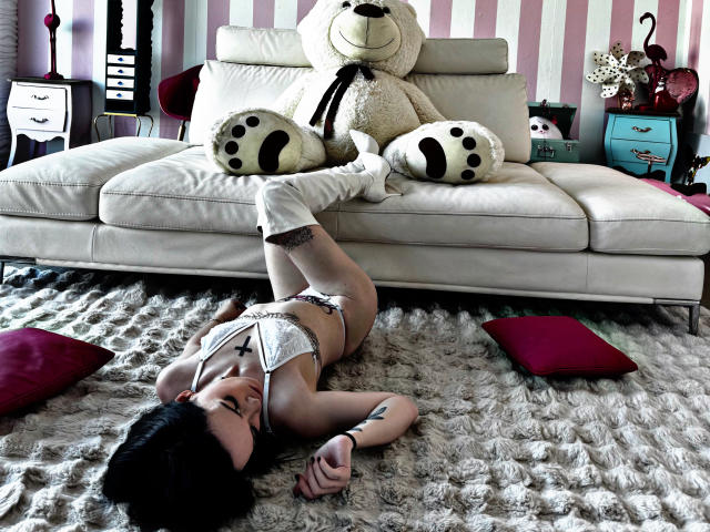 DreamArinna, lying on a fluffy rug, wears white lingerie and thigh-high socks, with one leg up on a white couch, next to a giant stuffed bear, showing her ass.