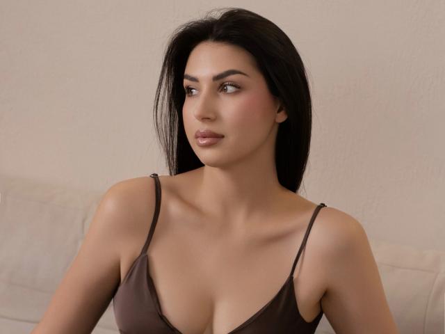 OliviaCluff has medium brown skin, long black hair, and is wearing a brown spaghetti-strap top, showing her tits and a subtle, sexy smile, looking to the side.