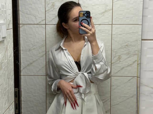 ColbyCupps with medium tits, wearing a white tied-up shirt, black lace bra, and white skirt, taking a mirror selfie with red nails, in a tiled bathroom.