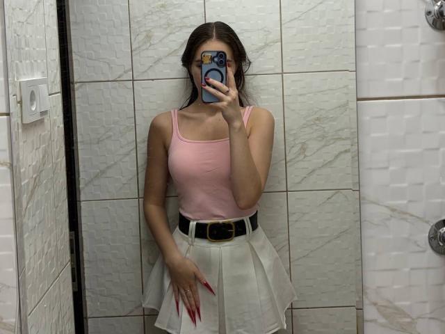 ColbyCupps with pink tank top, white pleated skirt, black belt, and red nails, holding phone covering face, mirror selfie, in tiled bathroom, showing tits and pussy hint.