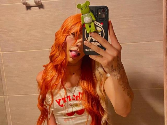 FloraWinx com cabelos longos laranja e loiros, língua de fora, vestindo uma camisa branca, segurando um celular com um adesivo de um sapo verde, braço tatuado, em um selfie no espelho do banheiro..