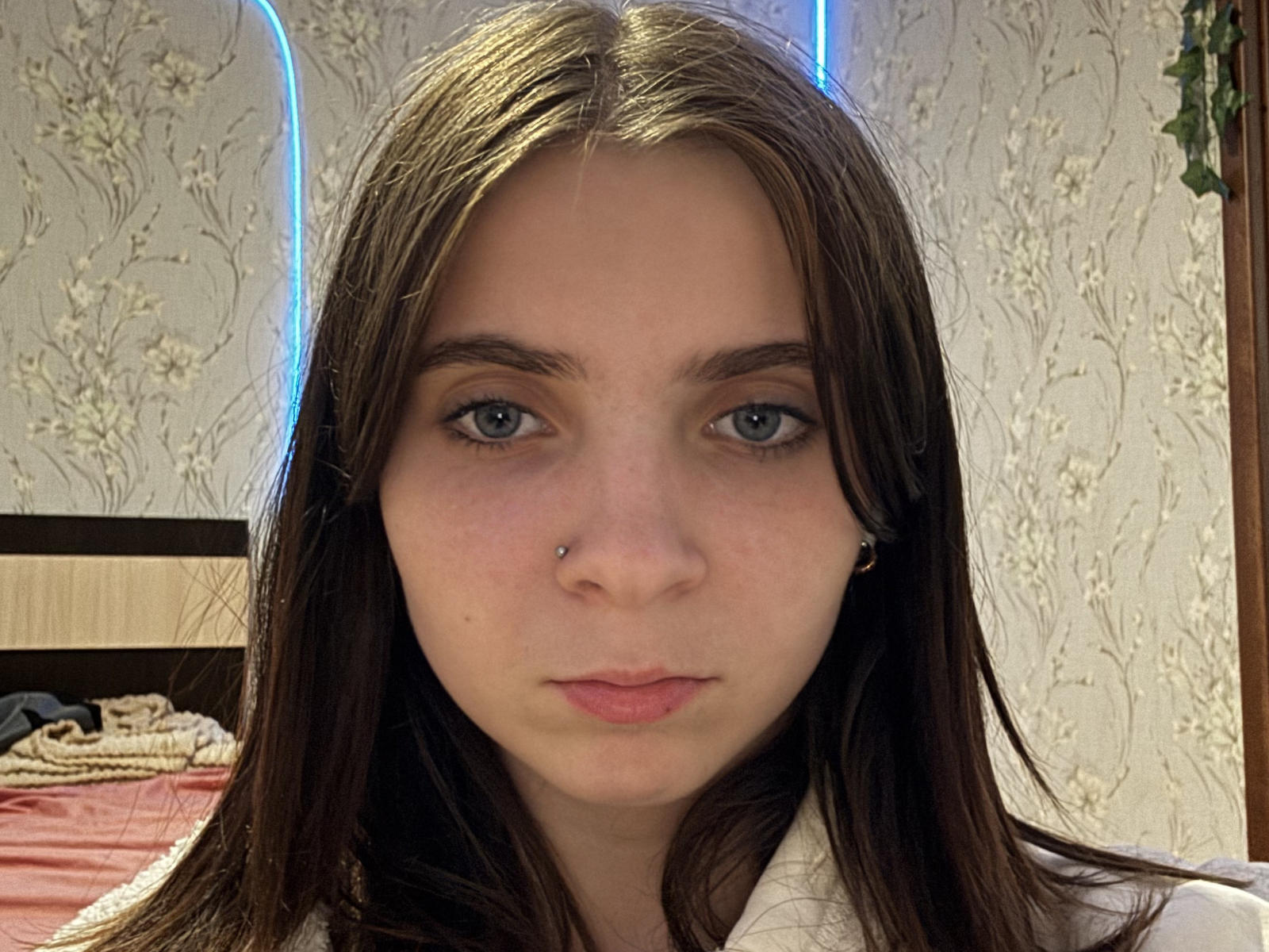 JacquiWinborn has straight brown hair, blue eyes, a nose piercing, and a serious expression. She's wearing a white shirt, with her ass barely visible in the background.