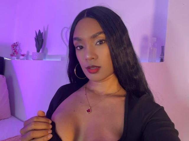NatLewis with long black hair, red lipstick, hoop earrings, and a black top showing cleavage, holding her top to reveal her tits. Purple lighting in the background.