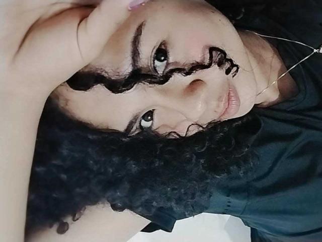 KirasPell with curly black hair, dark eyebrows, and a gentle smile, lying down, wearing a black shirt, with a silver necklace, looking up with a relaxed expression.