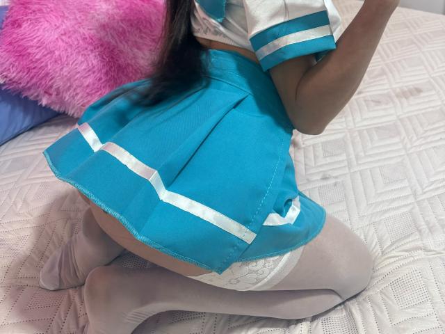 SammanthaOlssen on knees, wearing a blue and white schoolgirl outfit with white lace stockings, showing her ass. Pink fluffy pillow in the background.