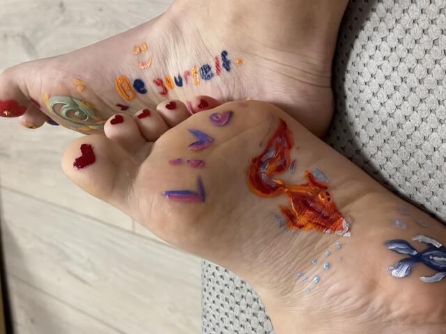 AlexXas' feet, painted with colorful, abstract designs on toes and soles, including a blue butterfly, red heart, orange flame, and green swirl, on a textured gray surface.