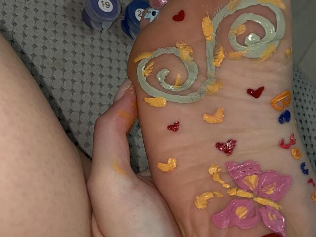 AlexXas's foot, painted with swirls, hearts, and a pink flower using yellow and red paint, held by AlexXas's hand. Background includes purple and blue bottles.