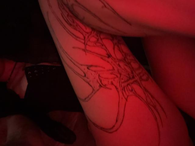 SimonneBrustkern's tattooed arm with intricate black ink designs, illuminated by red lighting. Visible black boot with studs on the left. Red light casts a sensual glow.