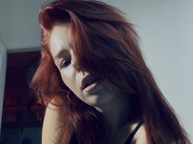 AlexXas has messy red hair covering one eye, subtle freckles on her face, and a slightly parted mouth with hoop earrings. She's looking down with a sexy, soft expression.