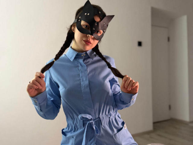 LeaMike with braided black hair, blue button-up dress, and black cat mask, fists up, showing her tits, in a bright room, wearing a playful, sexy expression.