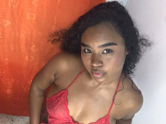 RoxxyEbony, met krullend zwart haar, donkerbruine huid en een rode kantbeha, kijkt omhoog met een lichte frons, waarbij haar kleine borsten en tatoeage op haar rechterarm zichtbaar zijn..
